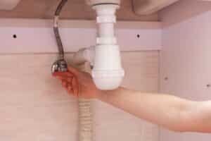 A person's hand loosens a pipe under a sink.